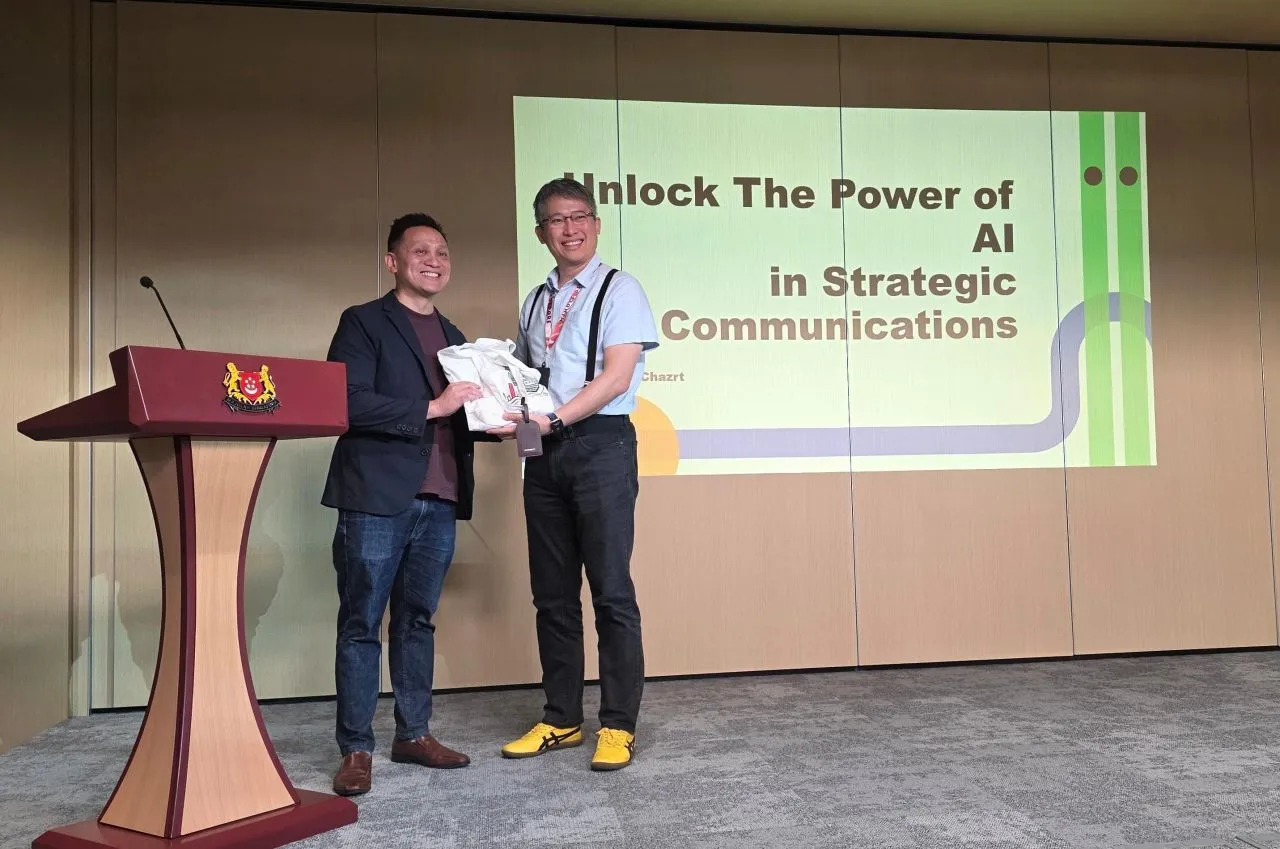 Roy Tan receiving a token of thanks on stage at MINDEF, in front of the slide Unlock the Power of AI in Strategic Communications